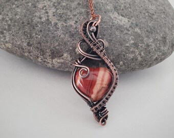 May include: A heart-shaped red and cream striped stone pendant encased in intricate, dark copper wire wrapping. The pendant features a swirling, decorative metal design that extends upward to a small loop, attached to a thin copper chain.