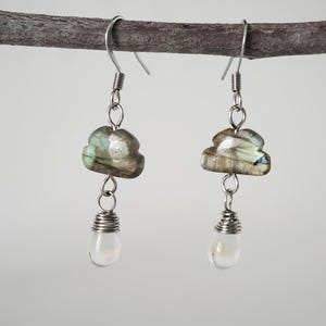 May include: A pair of silver-toned earrings. Each earring features a cloud-shaped, iridescent, gray-green stone, suspended below a wire-wrapped, pearlescent, teardrop-shaped bead. The earrings hang from a simple hook.