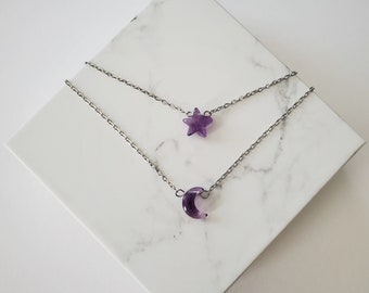 May include: Two layered necklaces with celestial pendants. The top necklace features a purple star pendant, and the bottom necklace has a purple crescent moon pendant. Both pendants are attached to a silver chain. The necklaces are displayed on a white marble surface.