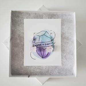 May include: A pair of light pink gemstone earrings with silver wire detailing, presented in a white gift box with a marble-like pattern. The earrings are displayed on a card with the text "ONE + SWIRL" and a geometric design.