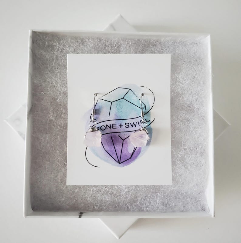 May include: A pair of light pink gemstone earrings with silver wire detailing, presented in a white gift box with a marble-like pattern. The earrings are displayed on a card with the text "ONE + SWIRL" and a geometric design.