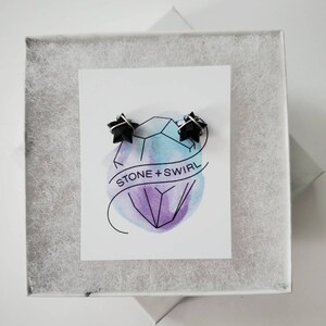 Puede incluir: Un par de pendientes negros en forma de estrella en una caja blanca con una tarjeta blanca que dice "STONE + SWIRL" y un dise&ntilde;o de acuarela de un cristal.