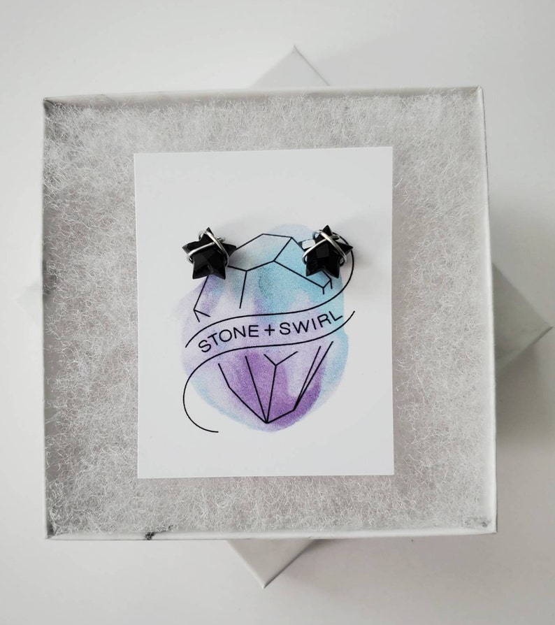 Puede incluir: Un par de pendientes negros en forma de estrella en una caja blanca con una tarjeta blanca que dice "STONE + SWIRL" y un dise&ntilde;o de acuarela de un cristal.