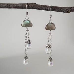 May include: A pair of handcrafted earrings suspended from a branch. Each earring features a cloud-shaped stone, a chain, and two iridescent, teardrop-shaped beads. The earrings are made with silver-colored metal.