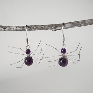 May include: A pair of silver spider earrings with amethyst gemstones. The spiders have eight legs and are made of wire.