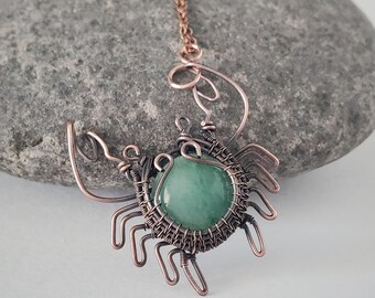 May include: A pendant necklace featuring a round, smooth, light green stone encased in dark copper wire wrapped to resemble a crab. The wire design includes two prominent claws and multiple legs extending from the central stone setting, suspended from a matching copper chain.