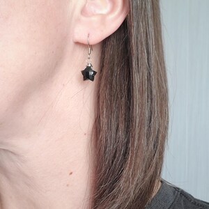 May include: A single black star-shaped earring with a silver hook is shown. The earring is hanging from the ear of a person. The star has multiple facets, giving it a textured appearance. The background is a light gray wall.