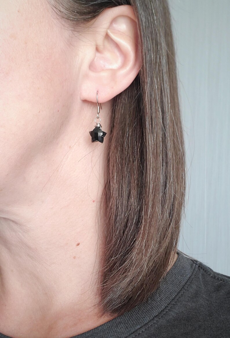 May include: A single black star-shaped earring with a silver hook is shown. The earring is hanging from the ear of a person. The star has multiple facets, giving it a textured appearance. The background is a light gray wall.