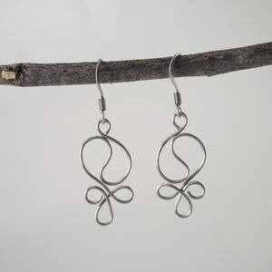 May include: A pair of silver-colored wire earrings with a unique, abstract design. Each earring features a looped top, a curved middle section, and a decorative bottom element. The earrings are suspended from a silver hook.