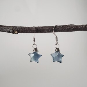 Op de afbeelding: Een paar hangende oorbellen met gefacetteerde, stervormige, doorschijnende blauwgrijze kralen. Elke ster is vastgezet met zilverkleurig draad en bevestigd aan een eenvoudige zilverkleurige metalen haak om aan de oorlel te hangen.