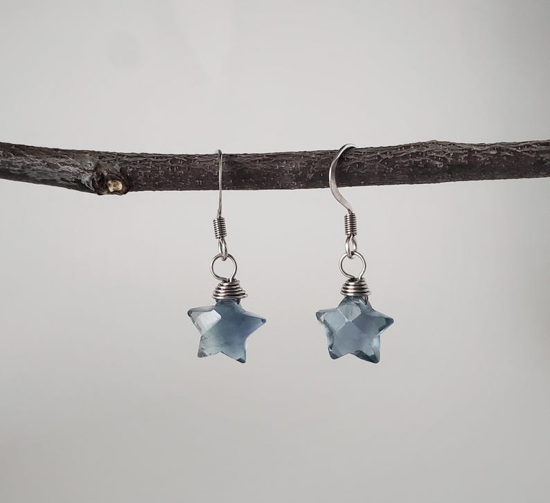 Op de afbeelding: Een paar hangende oorbellen met gefacetteerde, stervormige, doorschijnende blauwgrijze kralen. Elke ster is vastgezet met zilverkleurig draad en bevestigd aan een eenvoudige zilverkleurige metalen haak om aan de oorlel te hangen.