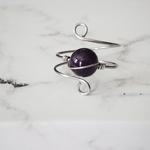 May include: A handcrafted ring featuring a polished, deep purple gemstone held within a swirling, silver-toned wire design. The ring's wirework has decorative loops at the ends, creating a unique and artistic piece of jewelry.