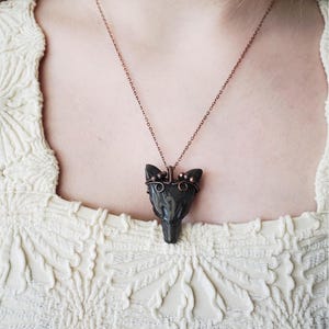 May include: A dark gray pendant necklace featuring a stylized wolf head. The pendant is suspended from a delicate copper-colored chain. The necklace is worn over a cream-colored, textured top.