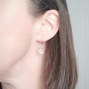 May include: A single earring with a small, light pink stone is visible. The stone is wire-wrapped and hangs from a silver-colored hook. The earring is worn on a person's ear, with the focus on the jewelry.