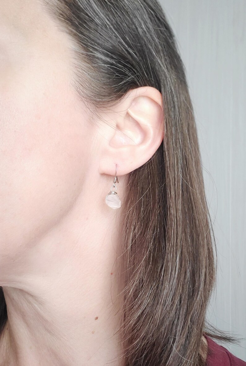 May include: A single earring with a small, light pink stone is visible. The stone is wire-wrapped and hangs from a silver-colored hook. The earring is worn on a person's ear, with the focus on the jewelry.