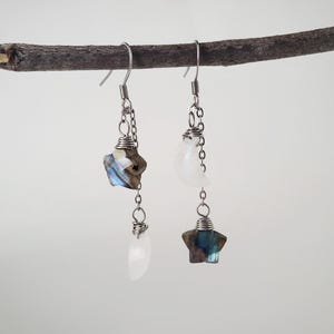 May include: A pair of silver earrings with celestial-themed charms. One earring features a faceted, blue-grey star and a white, teardrop-shaped stone. The other earring has a white moon and a faceted, blue-grey star. The earrings hang from a silver chain.