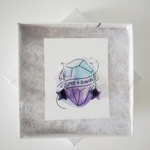 Op de afbeelding: Een paar stervormige oorbellen van paarse en witte steen met zilverkleurige metalen haakjes en draadwikkeling. De oorbellen worden gepresenteerd op een witte kaart met een aquarel kristalafbeelding en de tekst STONE + SWIRL in een witte doos.