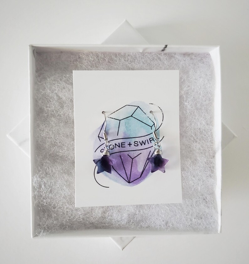 Op de afbeelding: Een paar stervormige oorbellen van paarse en witte steen met zilverkleurige metalen haakjes en draadwikkeling. De oorbellen worden gepresenteerd op een witte kaart met een aquarel kristalafbeelding en de tekst STONE + SWIRL in een witte doos.