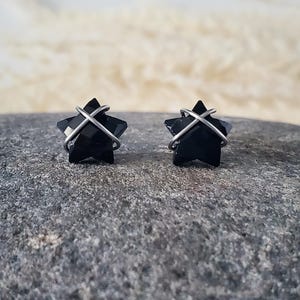 Puede incluir: Un par de pendientes de estrella negra con alambre plateado envuelto alrededor. Los pendientes est&aacute;n sobre una superficie de piedra gris.