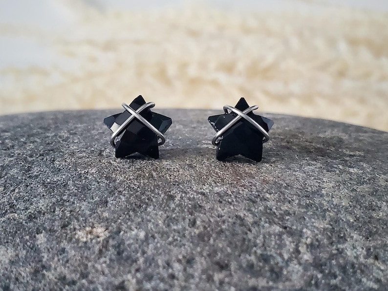 Puede incluir: Un par de pendientes de estrella negra con alambre plateado envuelto alrededor. Los pendientes est&aacute;n sobre una superficie de piedra gris.