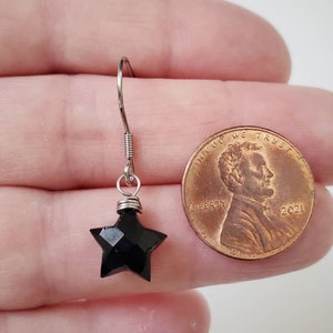 May include: A single black star-shaped earring with a silver hook. The earring is next to a penny for size comparison. The star has multiple facets, giving it a sparkling appearance. The earring is likely for pierced ears.