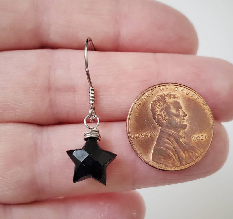May include: A single black star-shaped earring with a silver hook. The earring is next to a penny for size comparison. The star has multiple facets, giving it a sparkling appearance. The earring is likely for pierced ears.
