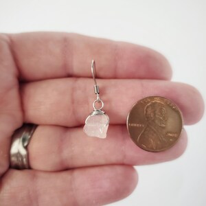 May include: A single, small, light pink raw stone earring with a silver wire wrap and hook. A penny is next to the earring for size comparison. The earring is held in a hand against a white background.