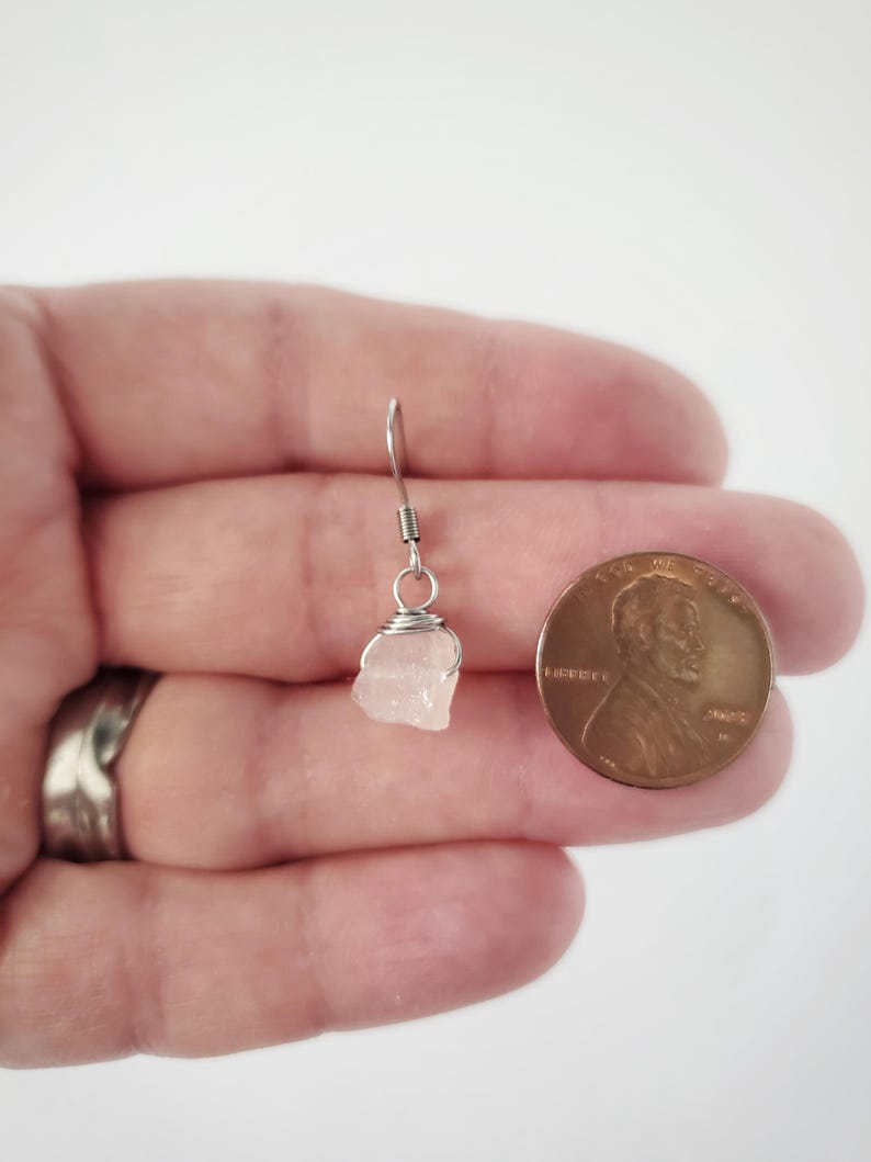 May include: A single, small, light pink raw stone earring with a silver wire wrap and hook. A penny is next to the earring for size comparison. The earring is held in a hand against a white background.