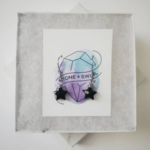 May include: A pair of black star-shaped earrings with silver-colored hooks, displayed in a white gift box. The earrings are on a card with the text "TONE + SWIRL" and a geometric design with blue and purple watercolor accents.