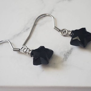 May include: A pair of dangle earrings featuring black star-shaped beads. The beads are attached to silver-colored fish hook ear wires. The earrings are displayed on a white marble surface.