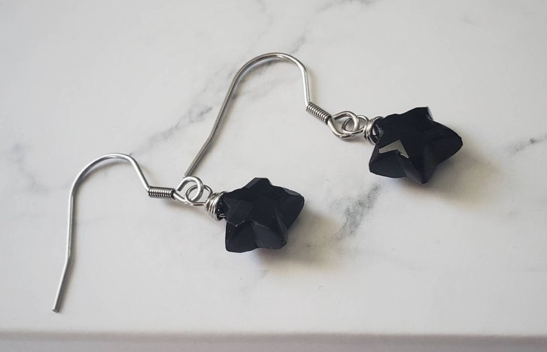 May include: A pair of dangle earrings featuring black star-shaped beads. The beads are attached to silver-colored fish hook ear wires. The earrings are displayed on a white marble surface.