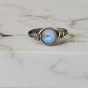 May include: A wire-wrapped ring with a round, iridescent moonstone. The ring has a silver-colored band and a circular setting that cradles the stone. The moonstone displays a blue and white color. The ring is set against a white marble surface.