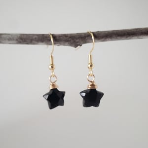 May include: A pair of star-shaped earrings. Each earring features a faceted black star pendant suspended from a gold-colored hook. The star is approximately 0.5 inches in diameter. The earrings are displayed against a neutral background.