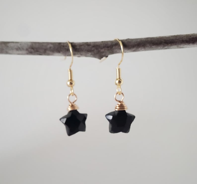 May include: A pair of star-shaped earrings. Each earring features a faceted black star pendant suspended from a gold-colored hook. The star is approximately 0.5 inches in diameter. The earrings are displayed against a neutral background.