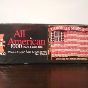 May include: A 1000-piece jigsaw puzzle box featuring an American flag made of Campbell's Soup cans. The box is black with red and white accents. The puzzle is titled "All American" and is 69 cm x 51 cm.