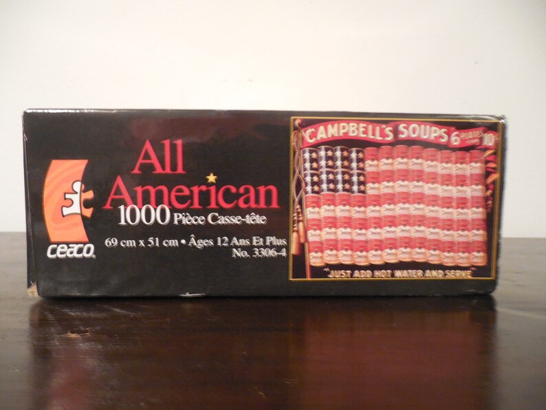 May include: A 1000-piece jigsaw puzzle box featuring an American flag made of Campbell's Soup cans. The box is black with red and white accents. The puzzle is titled "All American" and is 69 cm x 51 cm.