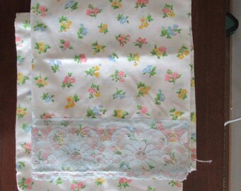 May include: White fabric with a floral pattern in pink, yellow, and blue. The fabric has a lace trim in a light blue and pink floral design. The fabric is folded over.