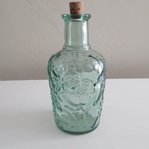 May include: A clear green glass bottle with a cork stopper. The bottle has a textured surface with a floral design.