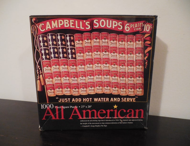 May include: A 1000-piece jigsaw puzzle box featuring a red, white, and blue American flag made of Campbell's Soup cans. The box reads "Campbell's Soups 6 Plates for 10c" and "Just Add Hot Water and Serve". The puzzle is titled "All American".