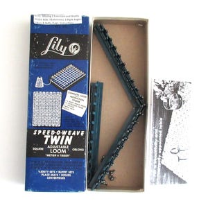 May include: A blue and white box with the text "Lily Speed-O-Weave Twin Adjustable Loom" and a picture of a loom. The box contains a blue plastic loom with metal pins. The loom is used for weaving.