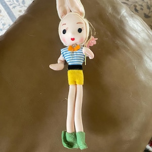 May include: A vintage doll with a bunny head, wearing a blue and white striped shirt, yellow shorts, and green boots. The doll has long, slender legs and is holding a small pink flower. The doll has a painted face with black eyes and a red mouth.
