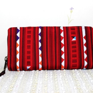 Puede incluir: Un monedero rectangular con un patrón de rayas rojas y blancas, una cremallera negra y un pompón rojo. El diseño presenta rayas verticales y detalles geométricos en naranja, azul y blanco. El fondo es una superficie blanca.
