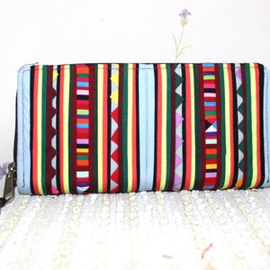 Puede incluir: Una cartera azul con cremallera con un patrón geométrico colorido. La cartera tiene un pompón verde unido al tirador de la cremallera.