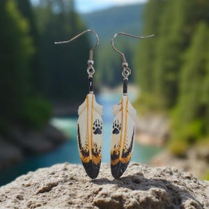 May include: A pair of dangle earrings featuring white feather-shaped pendants with orange and black markings. Each feather displays a black bear paw print design near the top and a dark black tip at the bottom, suspended from silver-toned metal hooks.