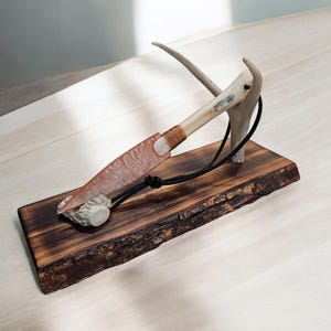 May include: A handmade knife with a bone handle and a dark brown wooden base. The knife is attached to the base with a leather cord and a bone antler.