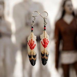 May include: A pair of feather earrings with a red handprint design. The earrings have a white base with black and gold accents at the bottom. The top of the earrings have a silver hook and a black and red beaded accent.