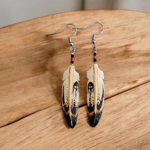 May include: A pair of feather earrings with silver hooks. The cream-colored feathers are adorned with black and red details, including a central design and accents along the edges. The earrings rest on a wooden surface.