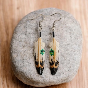 May include: A pair of dangle earrings featuring white feather-shaped charms with black and brown accents and green and black details. The earrings are hanging from silver hooks.