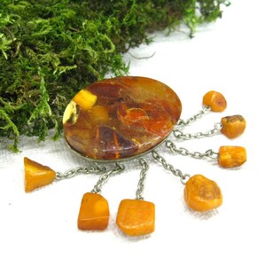 Puede incluir: Un broche vintage de &aacute;mbar con una piedra de &aacute;mbar ovalada grande rodeada de piedras de &aacute;mbar m&aacute;s peque&ntilde;as que cuelgan de cadenas plateadas.