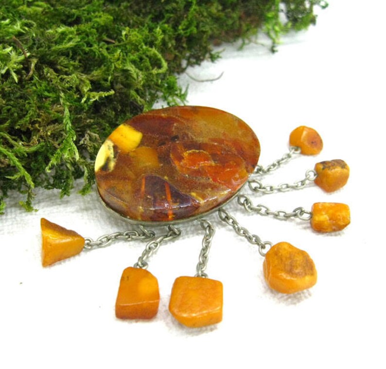 Puede incluir: Un broche vintage de &aacute;mbar con una piedra de &aacute;mbar ovalada grande rodeada de piedras de &aacute;mbar m&aacute;s peque&ntilde;as que cuelgan de cadenas plateadas.